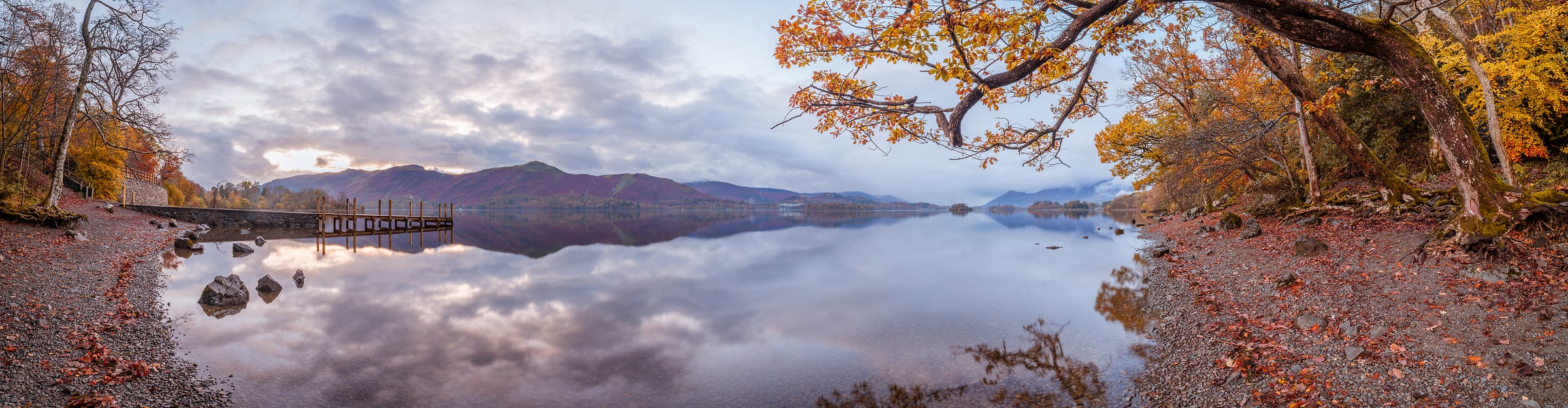 216 megapixels! A very high resolution, wall mural panorama photo of a calm lake at sunset with a pier, the rocky shoreline in the foreground, and mountains in the distance; panorama photograph created by Assaf Frank at Derwentwater Lake in Keswick, United Kingdom.